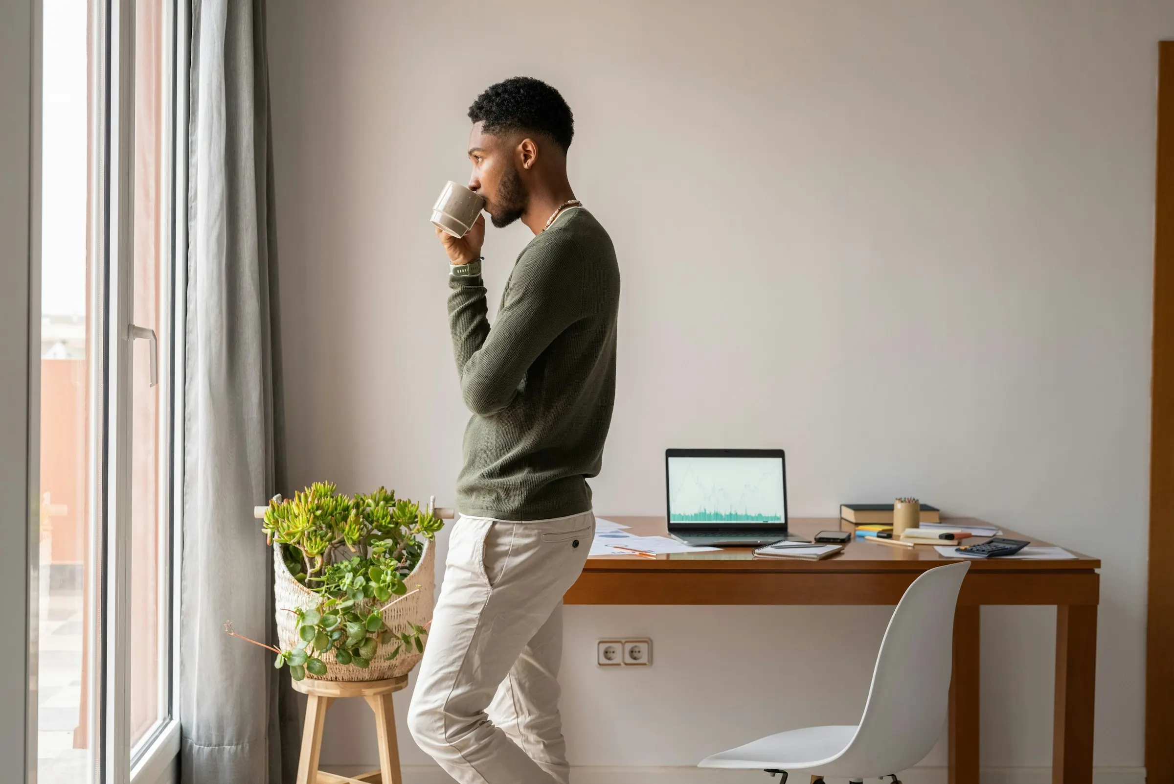 A developer drinking coffee by the window while his laptop works on the desk behind him